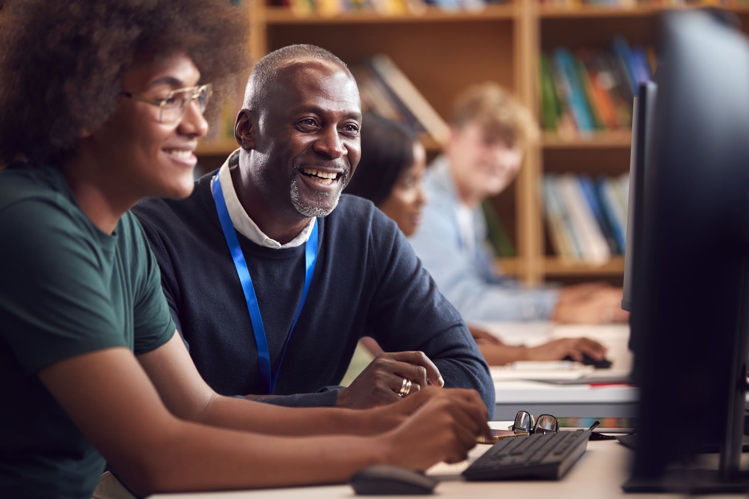 College Student Working At Computer In Library Being Helped By Tutor