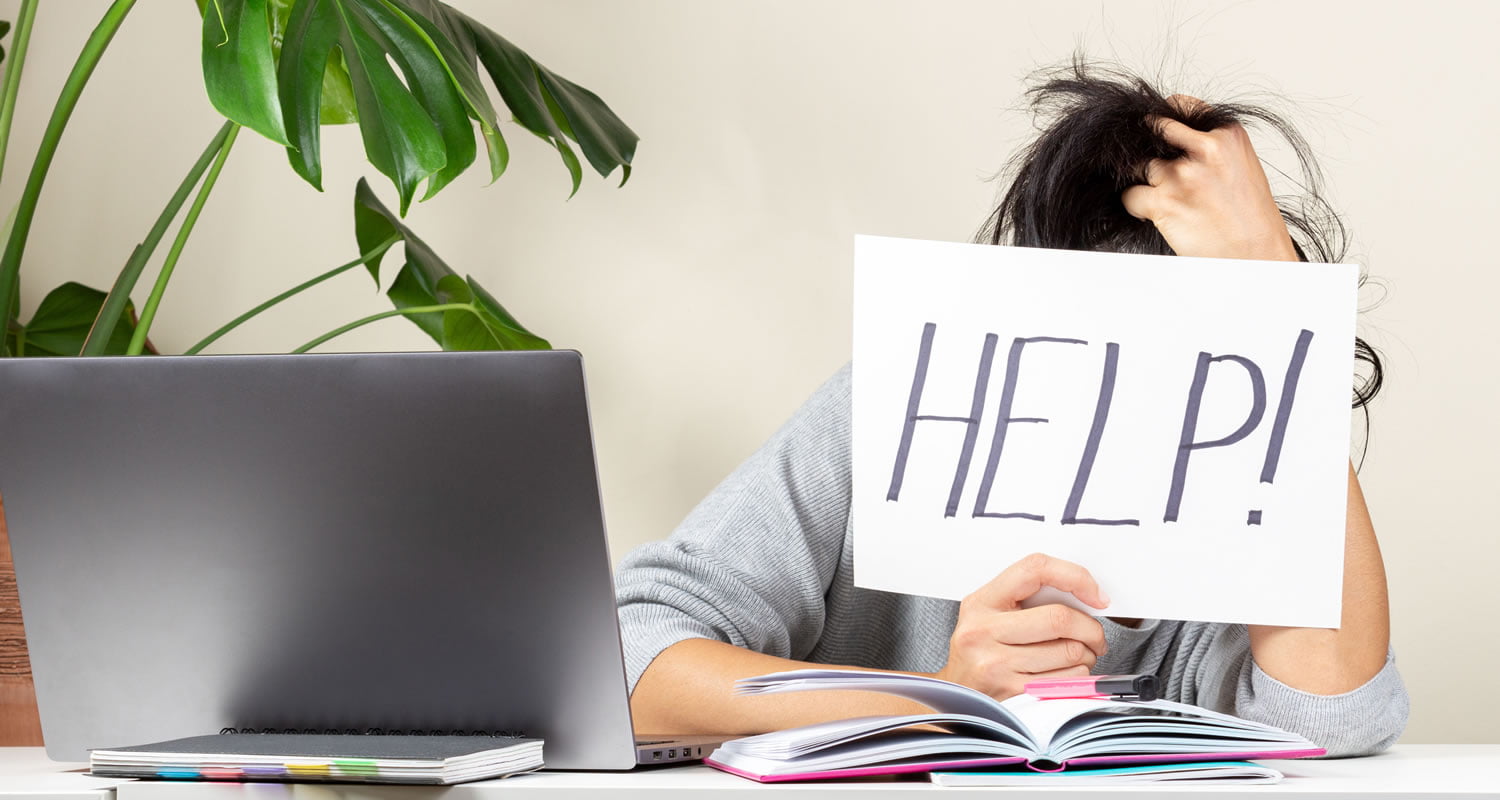 Tired frustrated student sitting at table and holding card with word Help.
