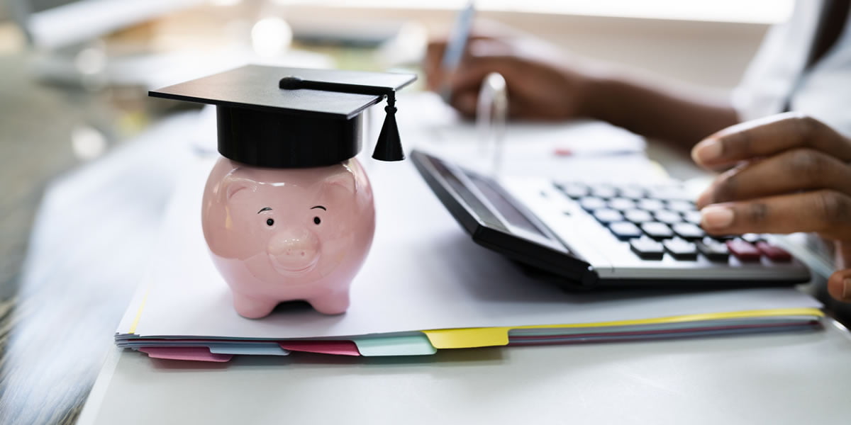 Piggy bank with graduation hat, sat on top of folders. Hands using calculator to work out cost